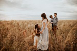 Pregnant woman in white lace dress stands with family in golden wheat field during sunset.
