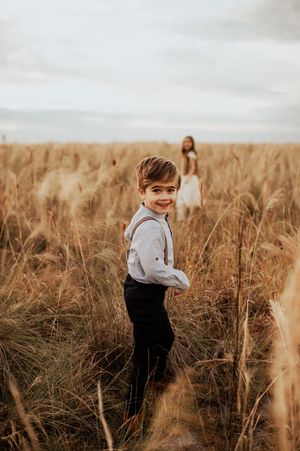 A young boy in suspenders smiles in a golden wheat field, with a girl in white dress standing in the background.