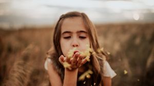 A young girl with closed eyes blowing golden confetti from her hands in a sunlit field.