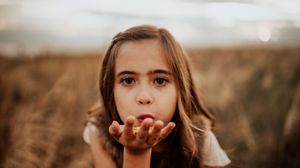 A young girl with long brown hair blowing a kiss outdoors in a golden field at sunset.