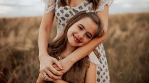 A woman hugs a smiling young girl from behind in a golden field, capturing a warm mother-daughter moment outdoors.