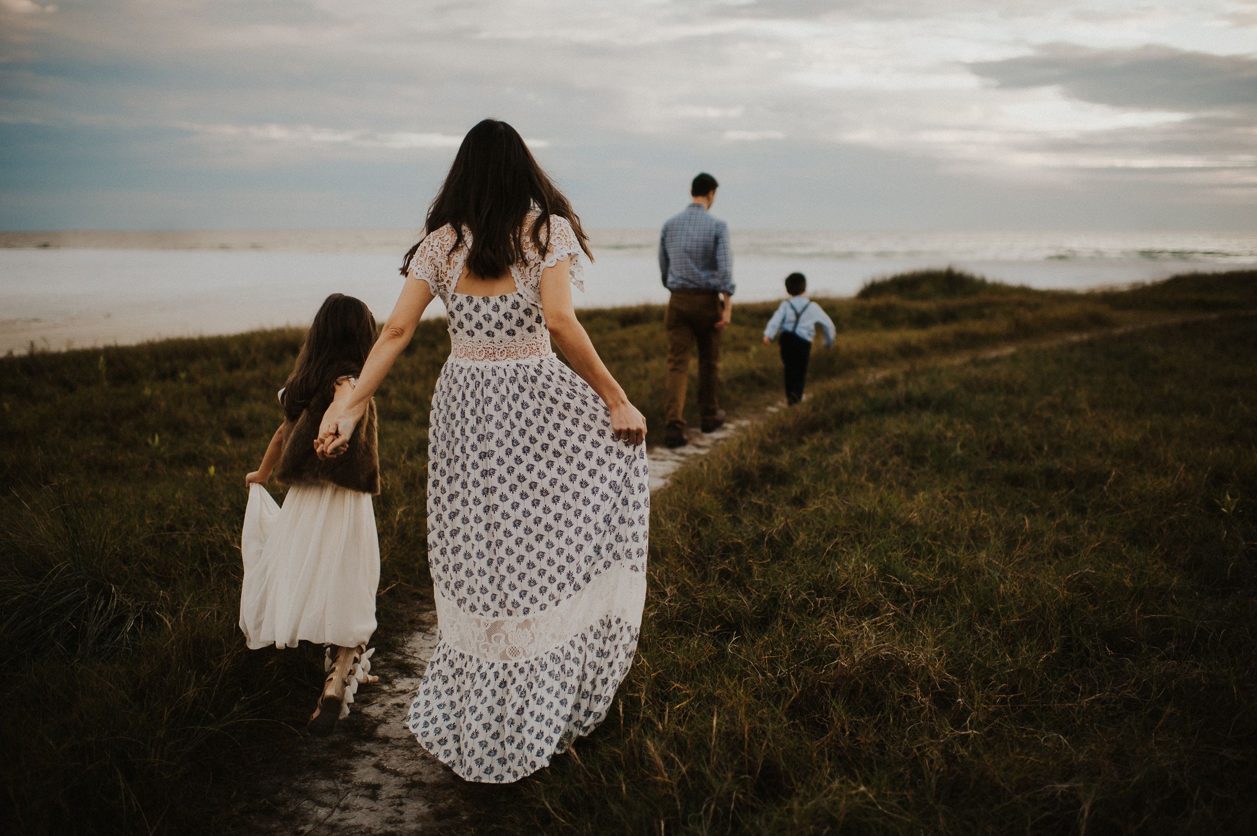 Family walking on coastal path at sunset, mother in floral dress holding daughter's hand, father and son ahead.