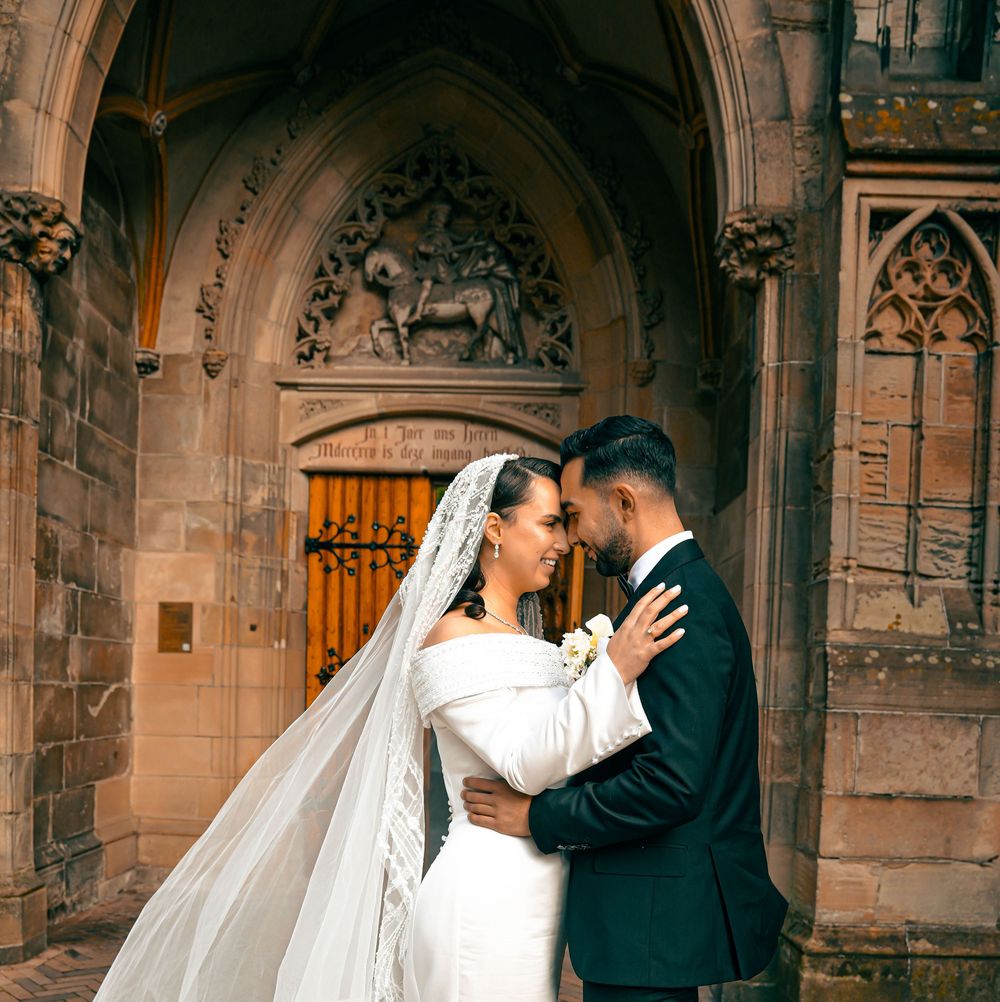Bride and groom embrace romantically outside a historic Gothic stone church, bride wearing a white gown and cathedral veil.