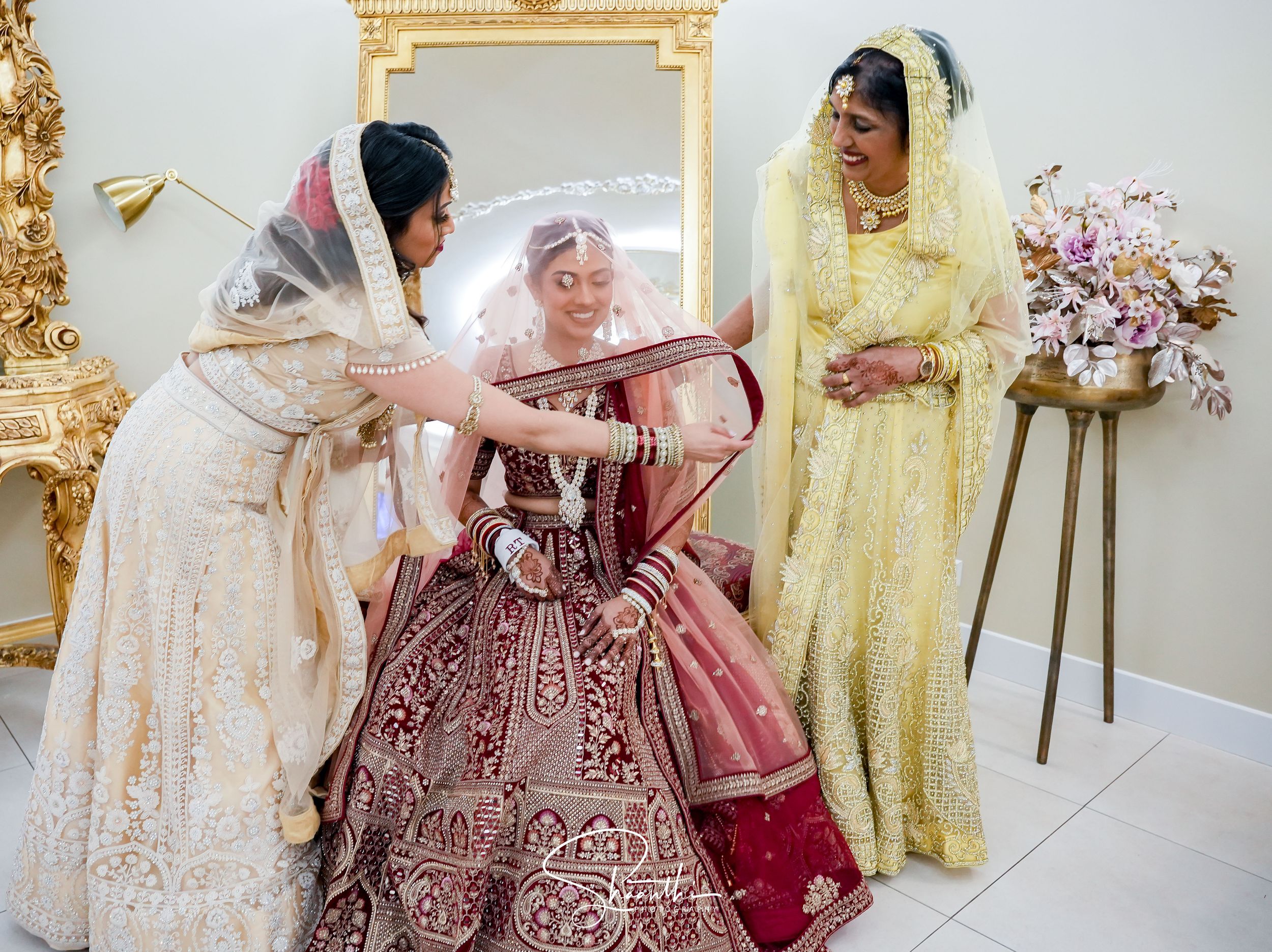 A group in traditional South Asian attire helps adjust an ornate burgundy and gold bridal lehenga during a wedding preparation.