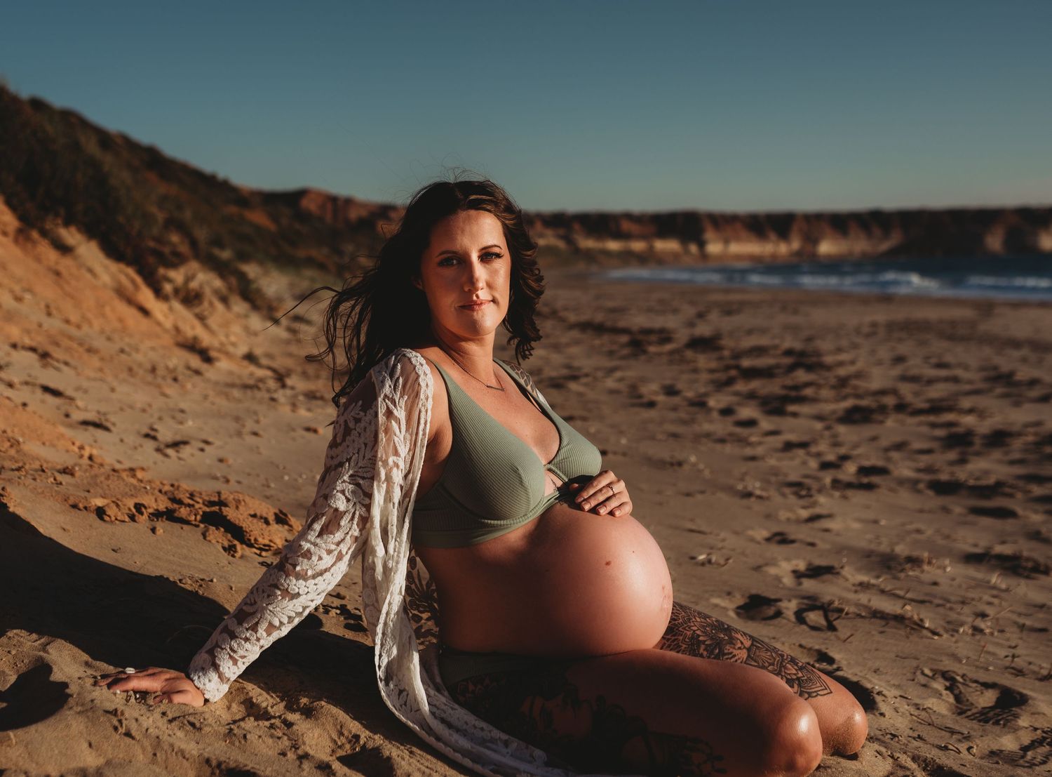 Expectant mother posing on sandy beach with cliffs in background at golden hour.