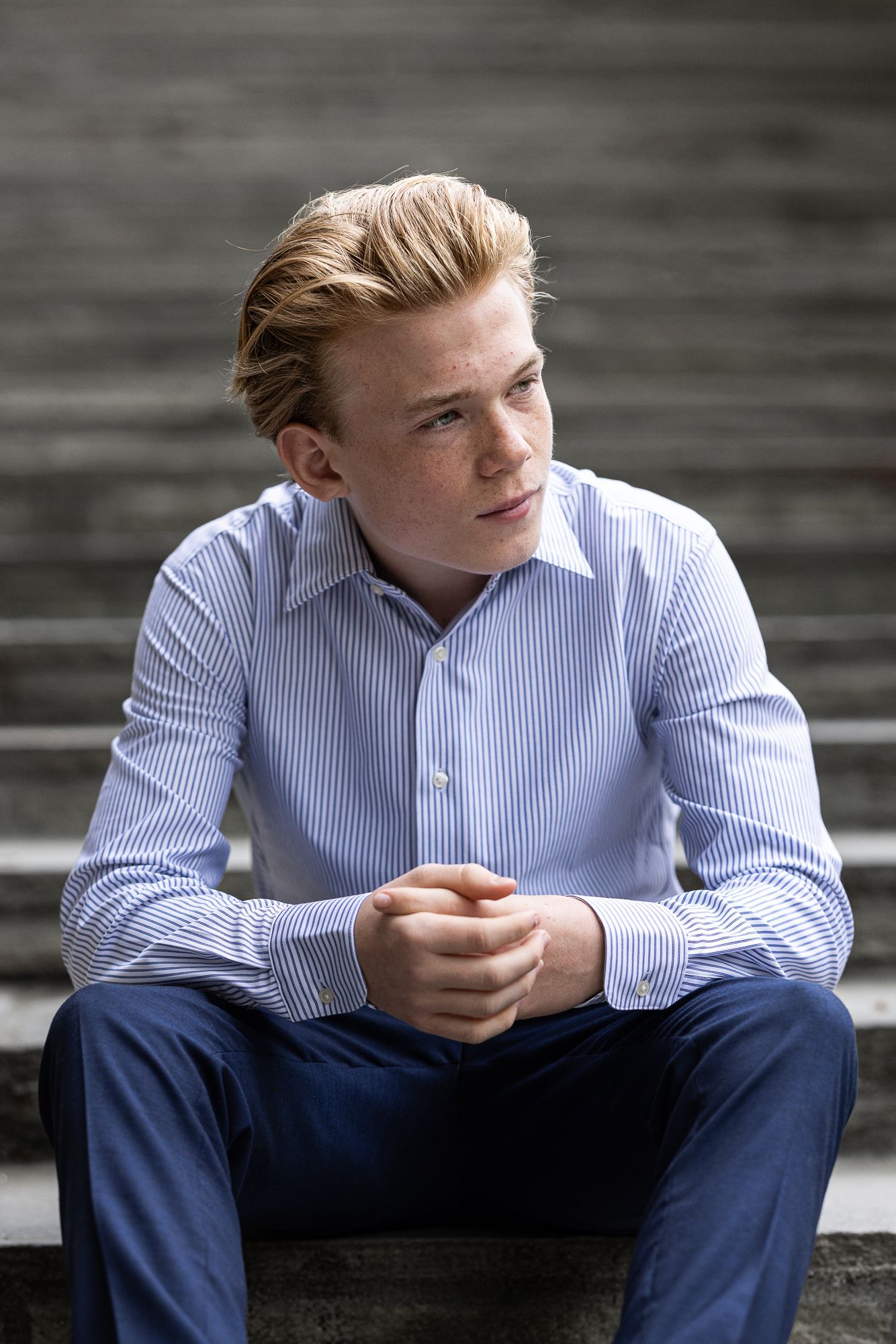 A person in a light blue striped dress shirt and navy pants sits thoughtfully on concrete steps.