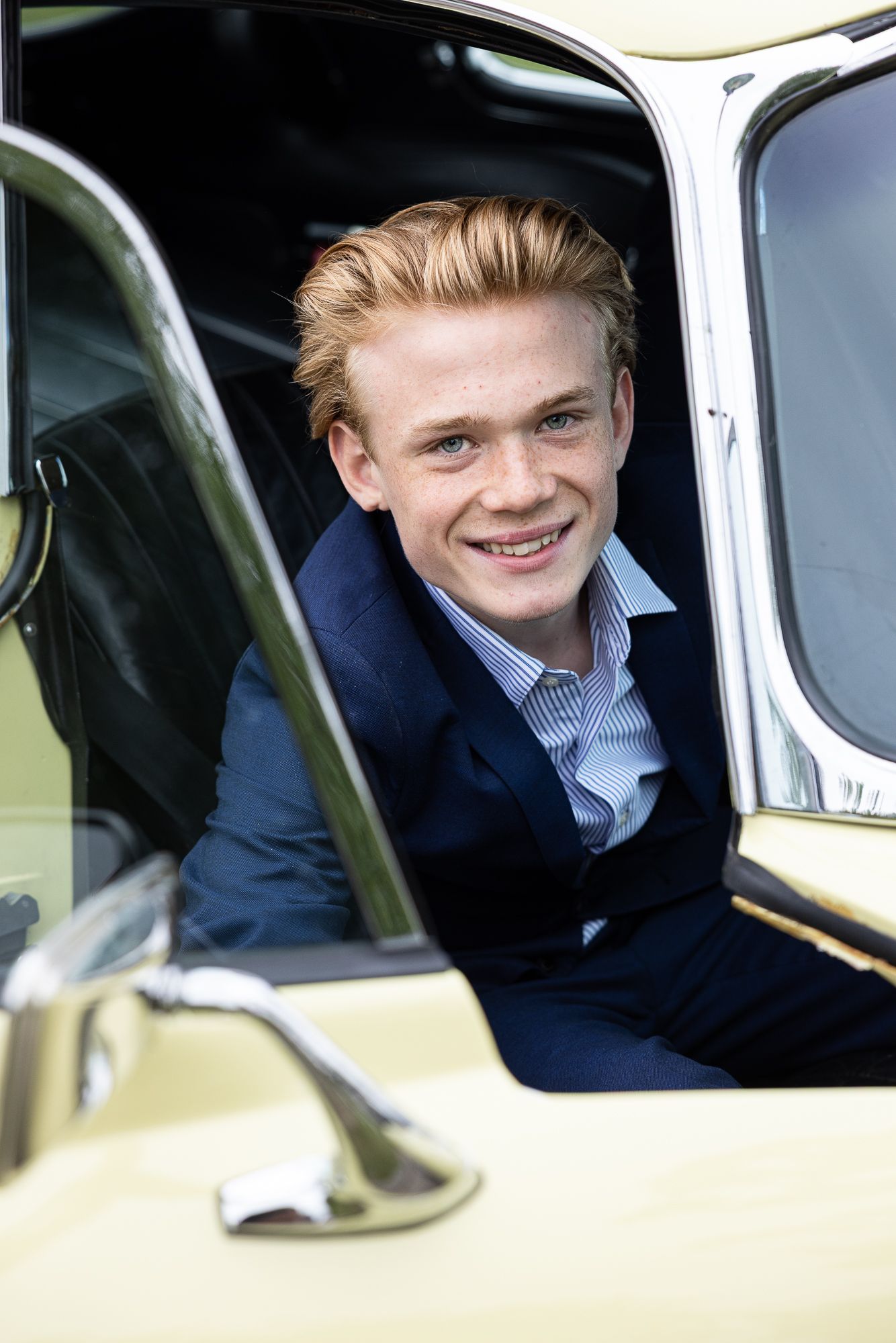 A smiling person sits in the driver's seat of a vintage car wearing a navy blazer and striped shirt.
