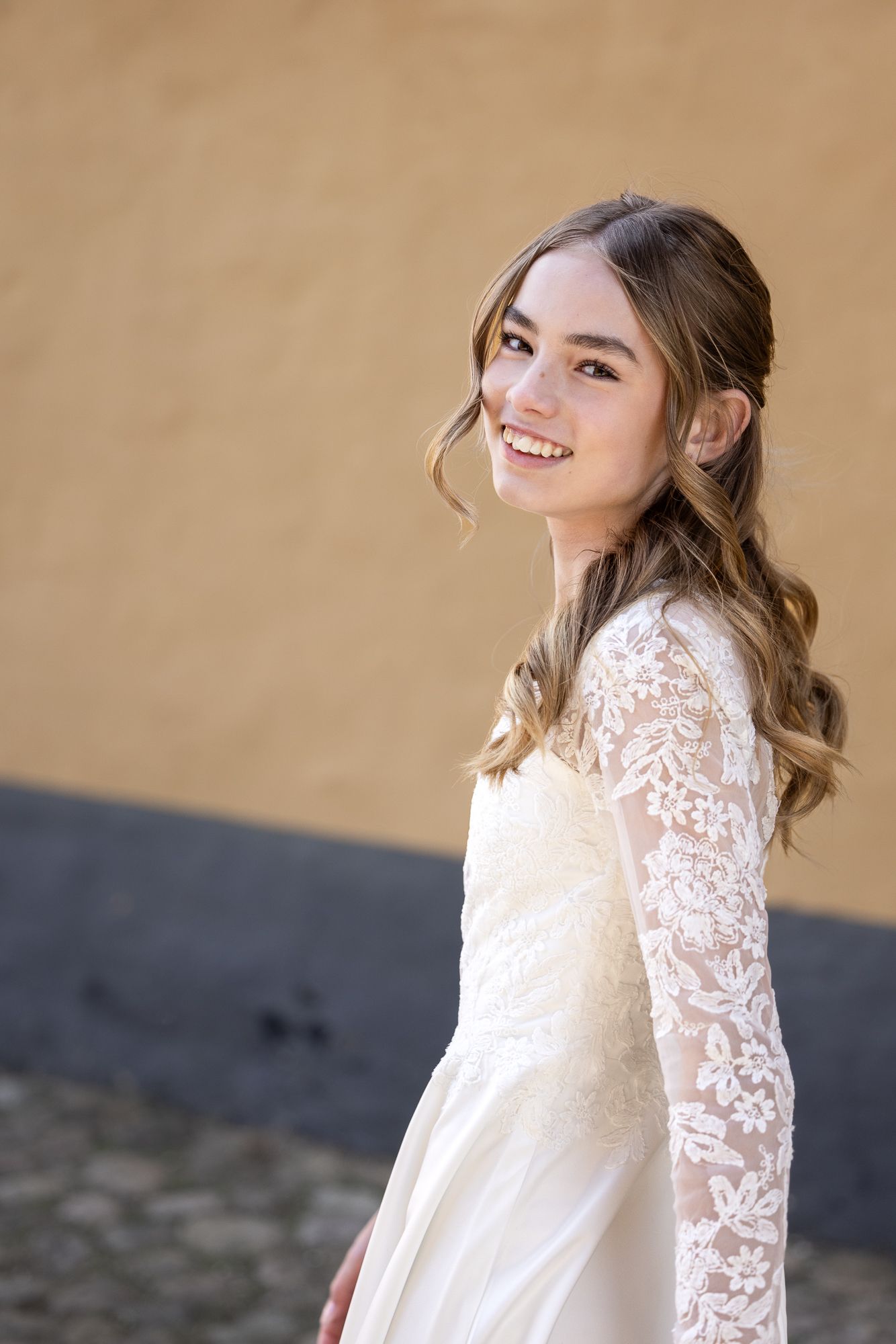 A smiling young woman in a white lace wedding dress with long sleeves poses against a tan and gray wall.