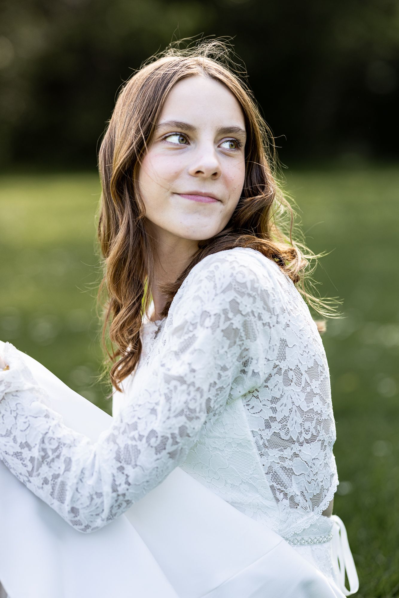 Young girl in white lace dress sitting outdoors in green park, smiling softly with long auburn hair.