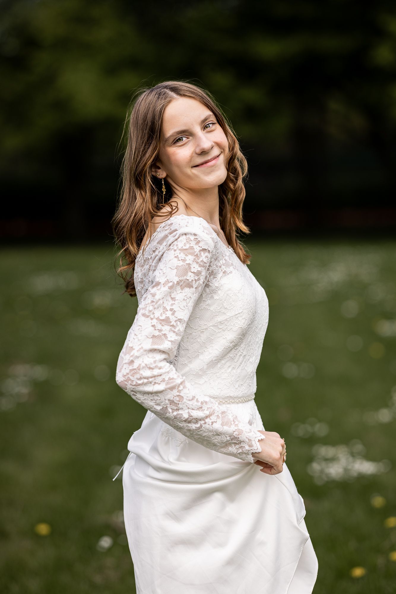 Young woman in white lace wedding dress smiling outdoors in green garden setting.