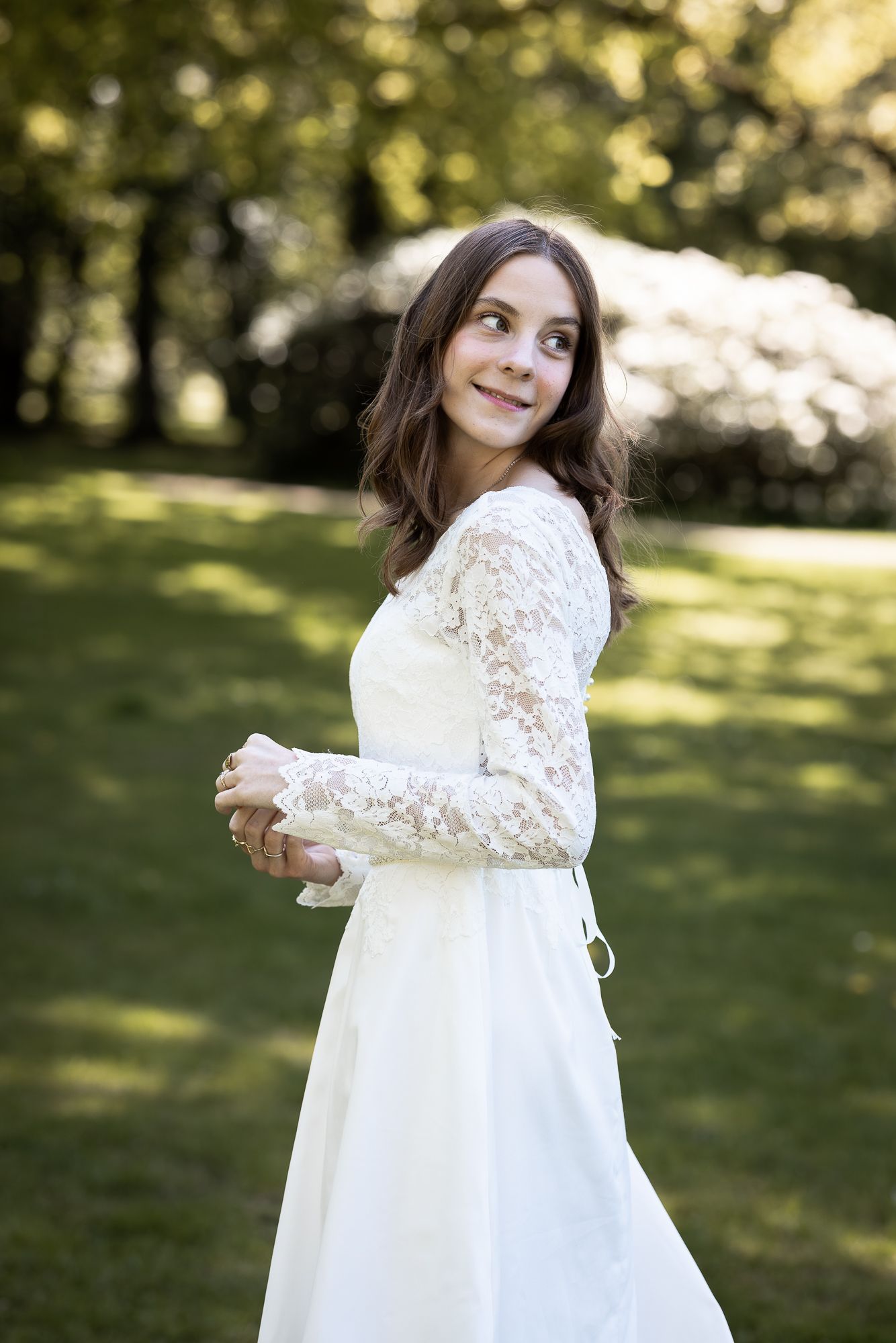Young woman in white lace wedding dress smiling over shoulder in lush green garden setting.