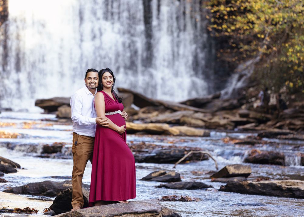 Expecting couple poses on rocky waterfall landscape, wearing red dress and white shirt.