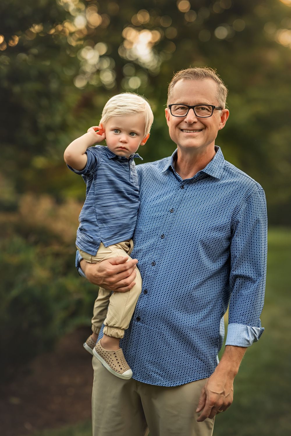 A heartwarming outdoor portrait shows a senior man in a blue checkered shirt holding a small child in casual clothing.