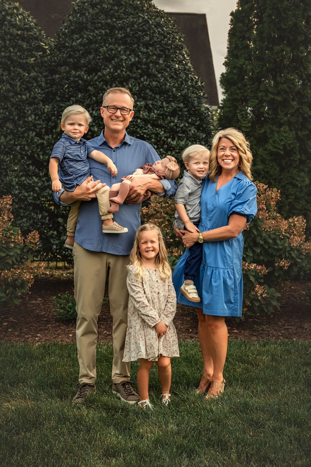 A family of five poses together outdoors in coordinated blue and neutral outfits against a green hedge background.