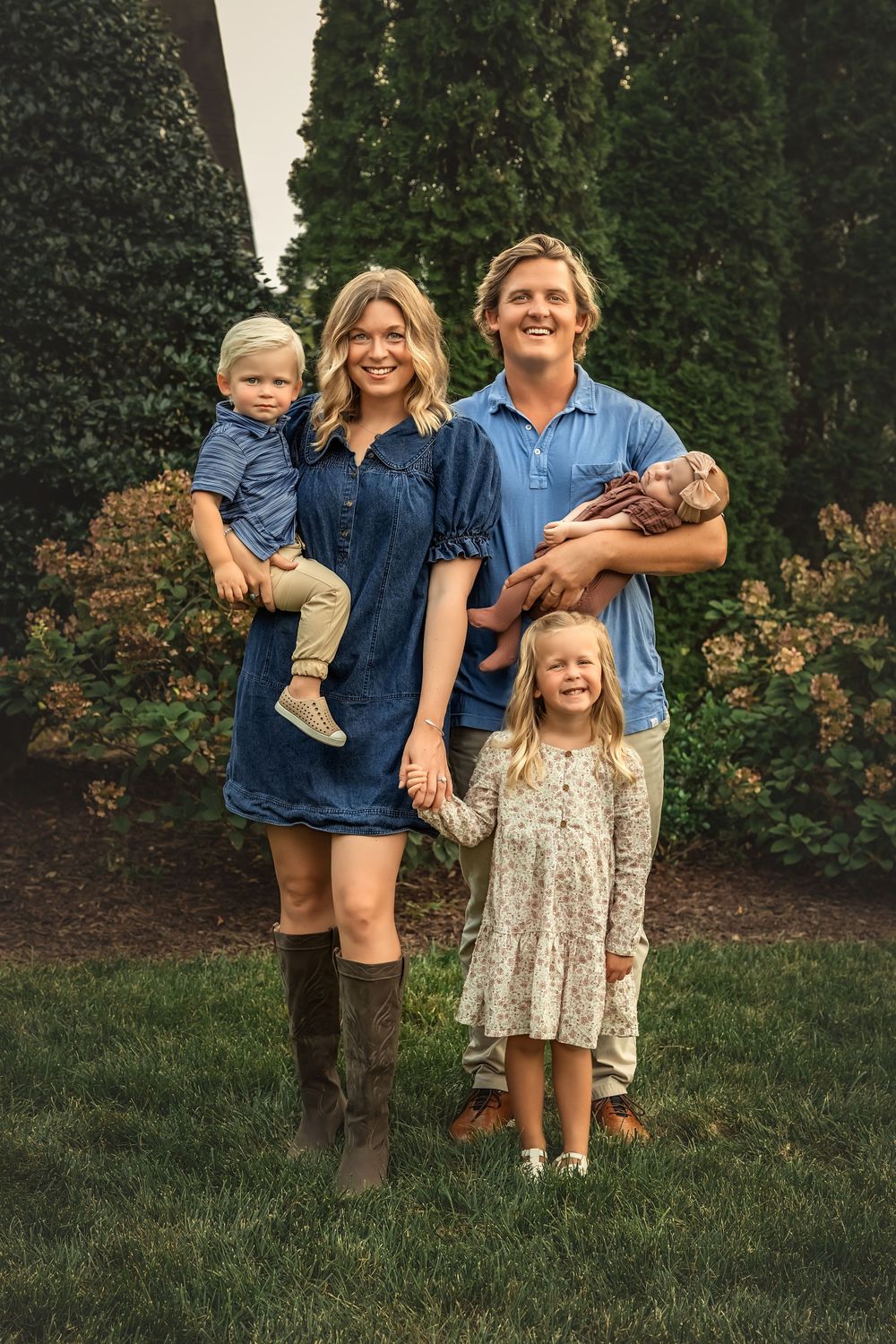 Family wearing coordinated denim and neutral outfits poses together in front of green hedges in outdoor garden setting.