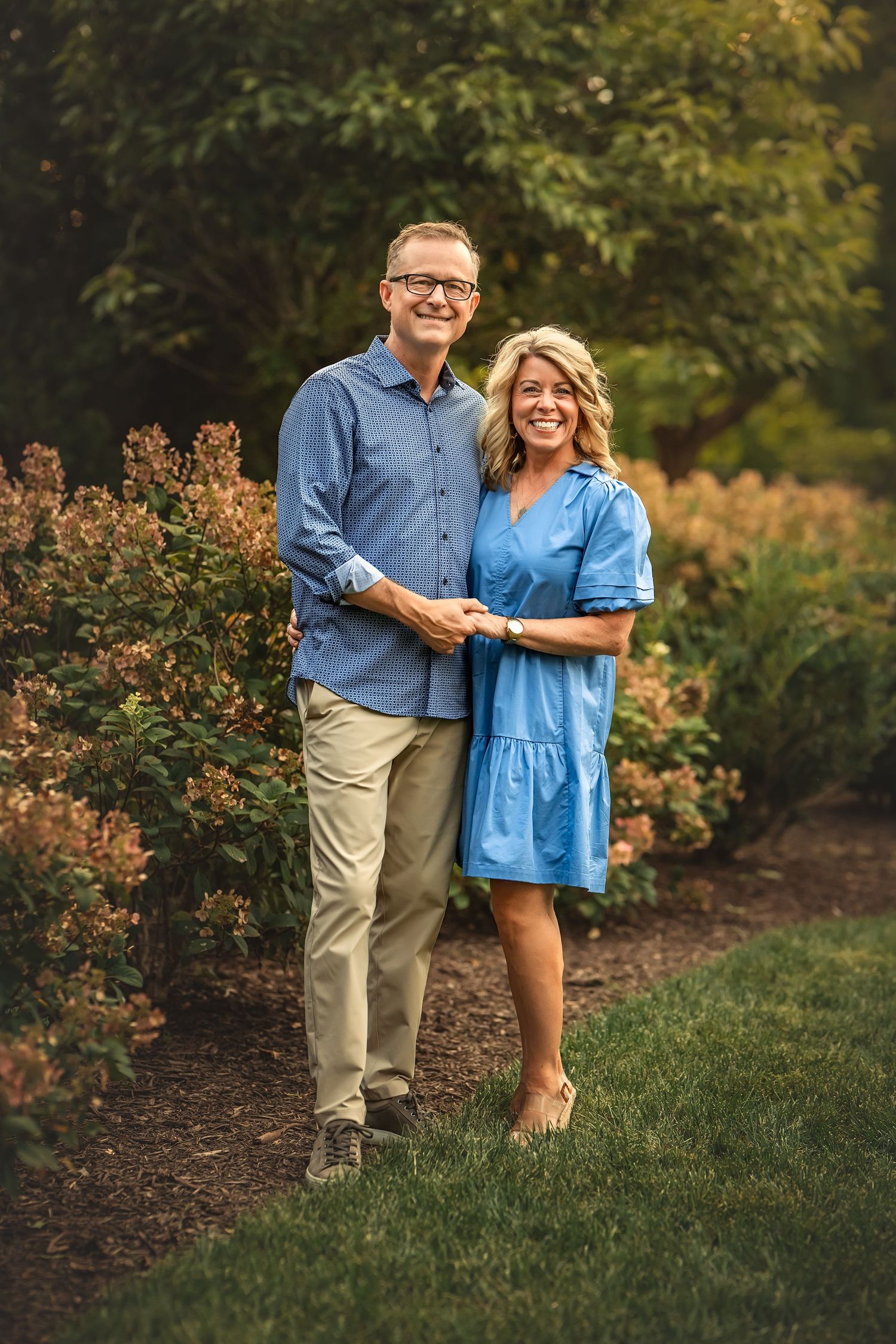 A couple wearing coordinating blue and khaki outfits embrace while standing in a garden with flowering shrubs at sunset.