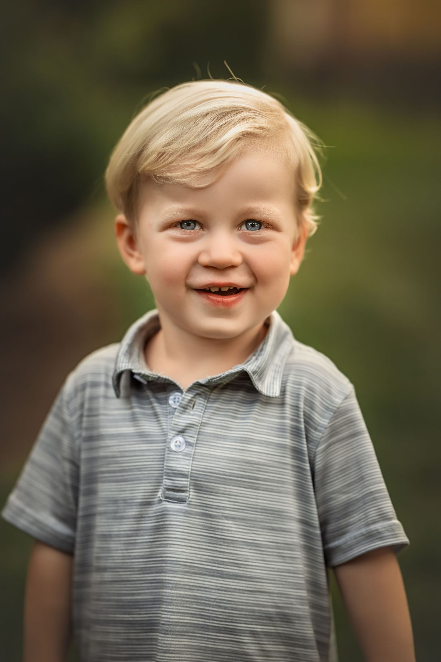 A young child in a gray striped polo shirt smiles brightly outdoors against a blurred natural background.