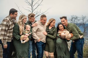 An extended family wearing plaid and olive green clothing laughs together while posing outdoors against a winter landscape.