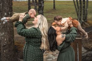 Two sisters in matching green dresses lifting their small babies outdoors in an autumn forest setting.