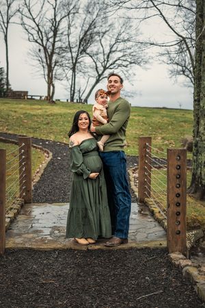 Family in coordinating green outfits poses together on wooden bridge during outdoor maternity photoshoot.