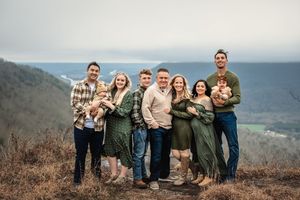 An extended family wearing coordinated green and tan outfits pose for a casual outdoor photo session against mountain scenery.