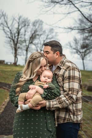 Family outdoors in autumn with baby embracing on a path near bare trees.