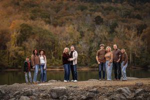 An extended family gathers for photos on a rocky outcrop by a serene lake during autumn with trees in the background.