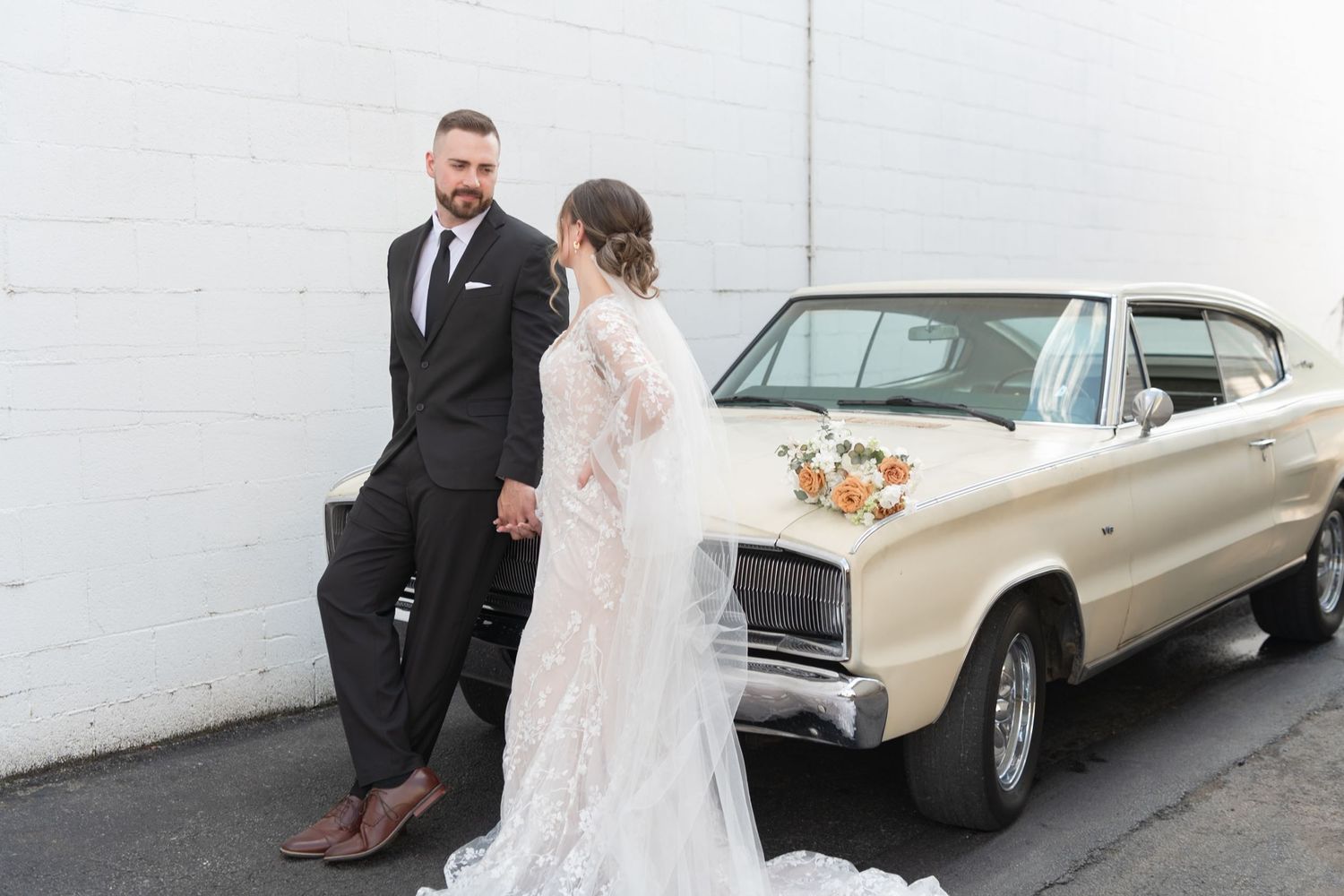 Wedding couple poses by vintage car against white wall.