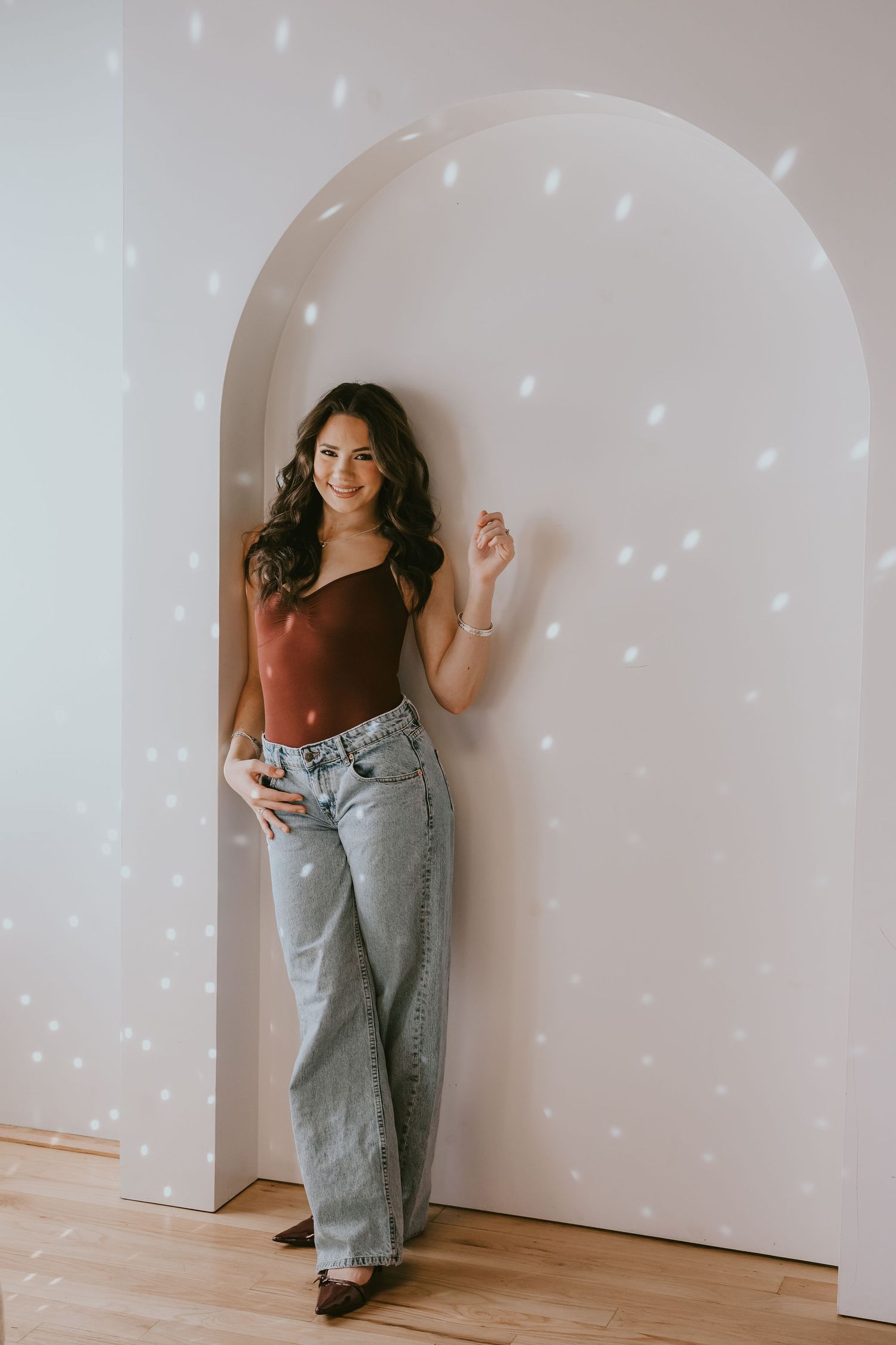 Casual portrait of woman in burgundy top and jeans against white arch with sparkle lights.