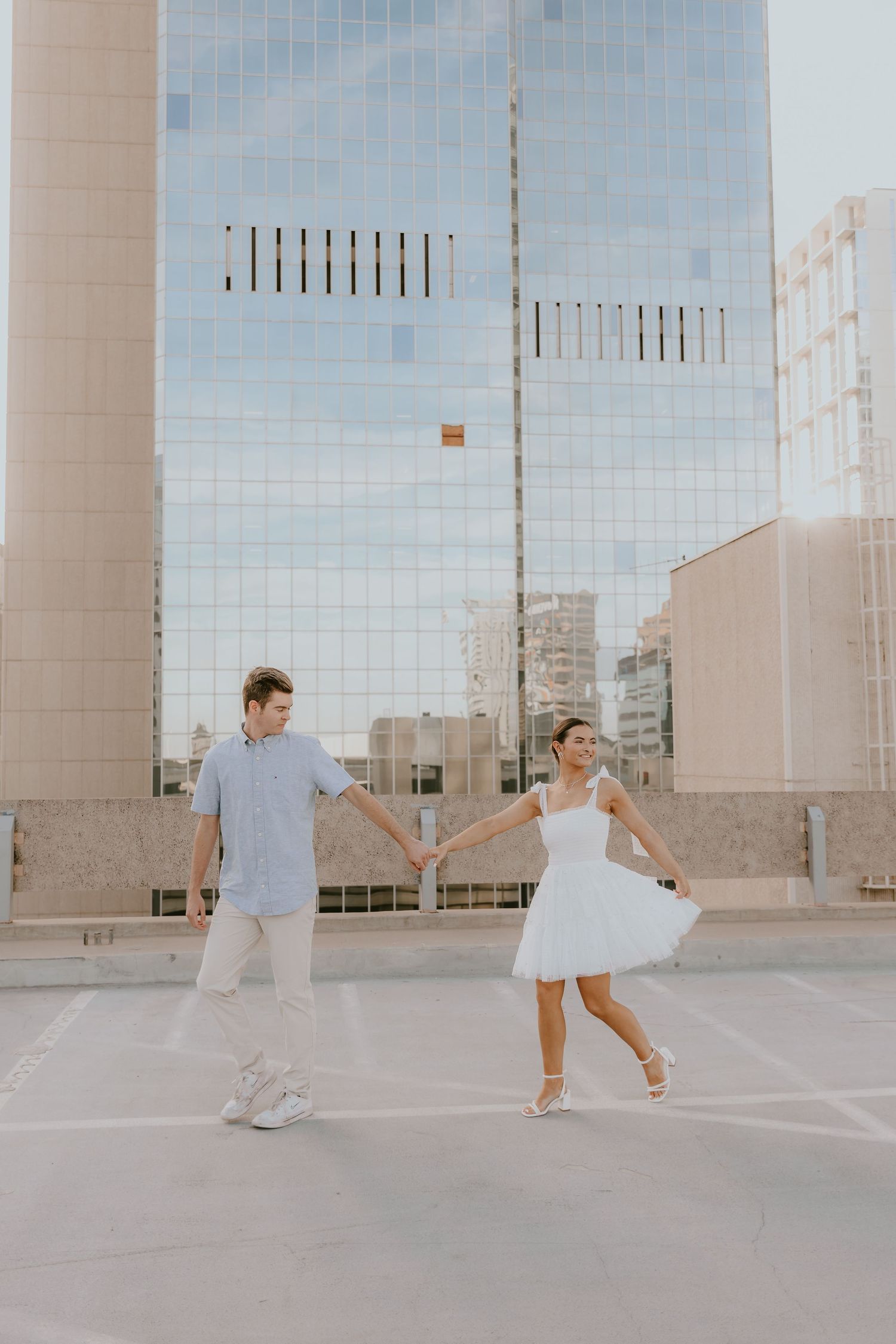 Couple in white outfits dancing on urban rooftop with glass building backdrop.