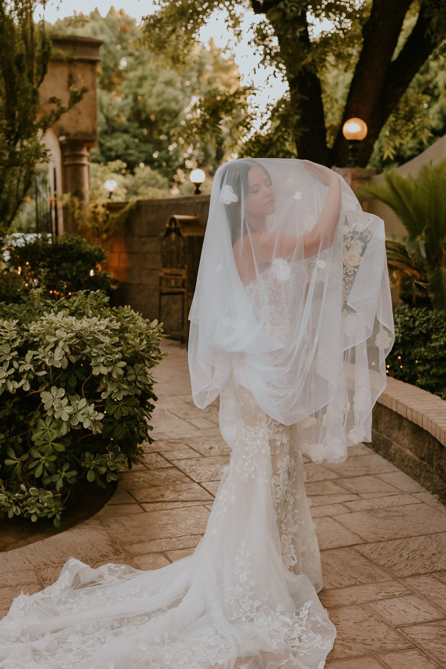 Bride in lace gown with flowing veil poses in garden setting.