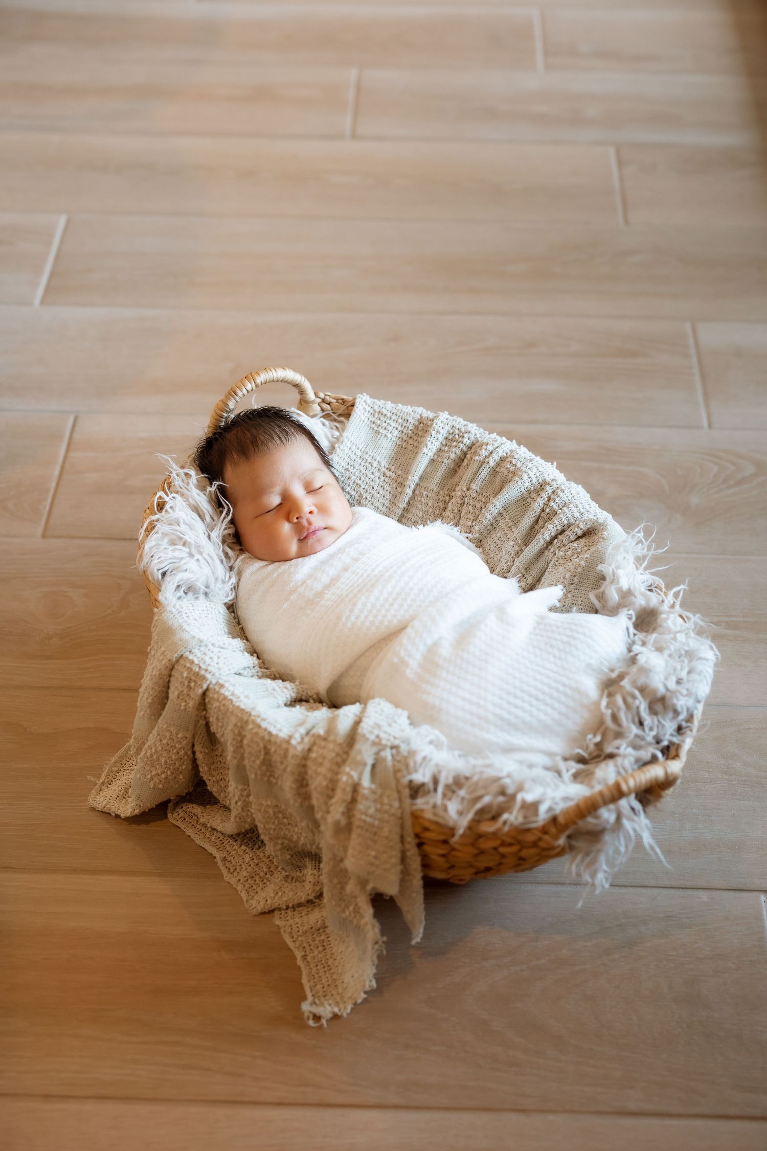 Newborn baby swaddled in white wrapped in cozy cream blankets on hardwood floor.
