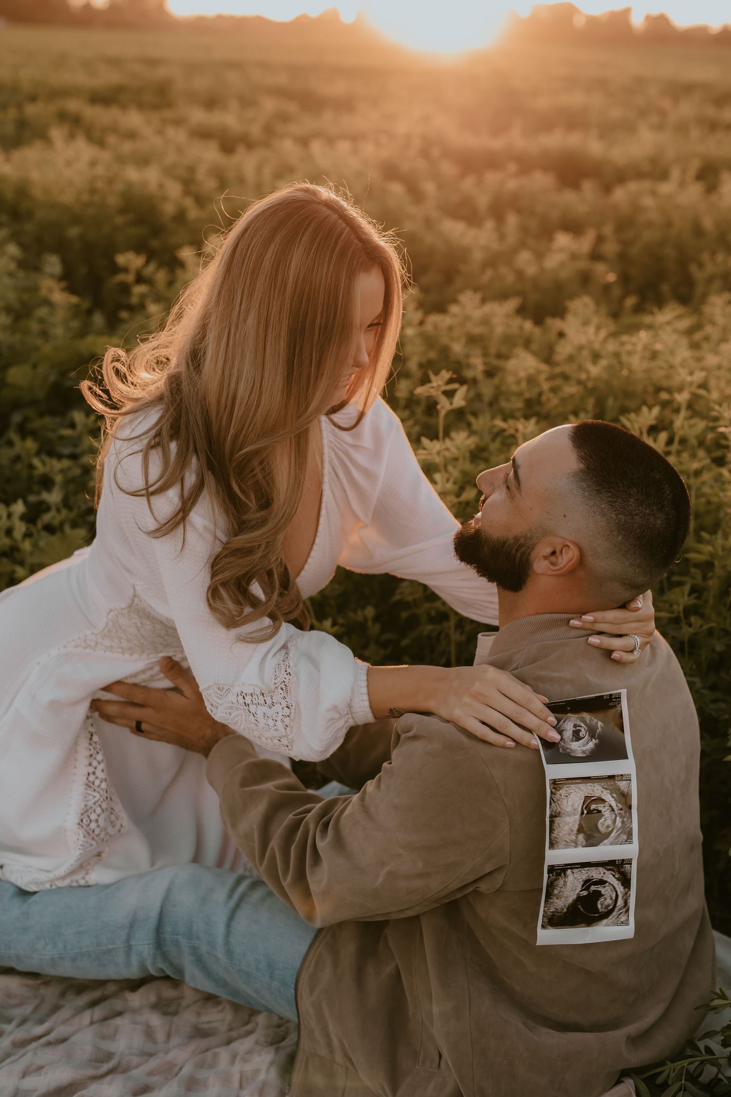 Couple embraces during sunset photoshoot in field with ultrasound photos.