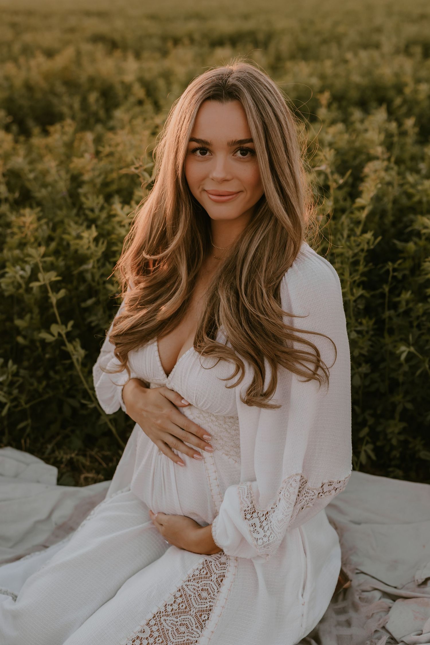 Woman in white lace dress poses during sunset maternity photoshoot.