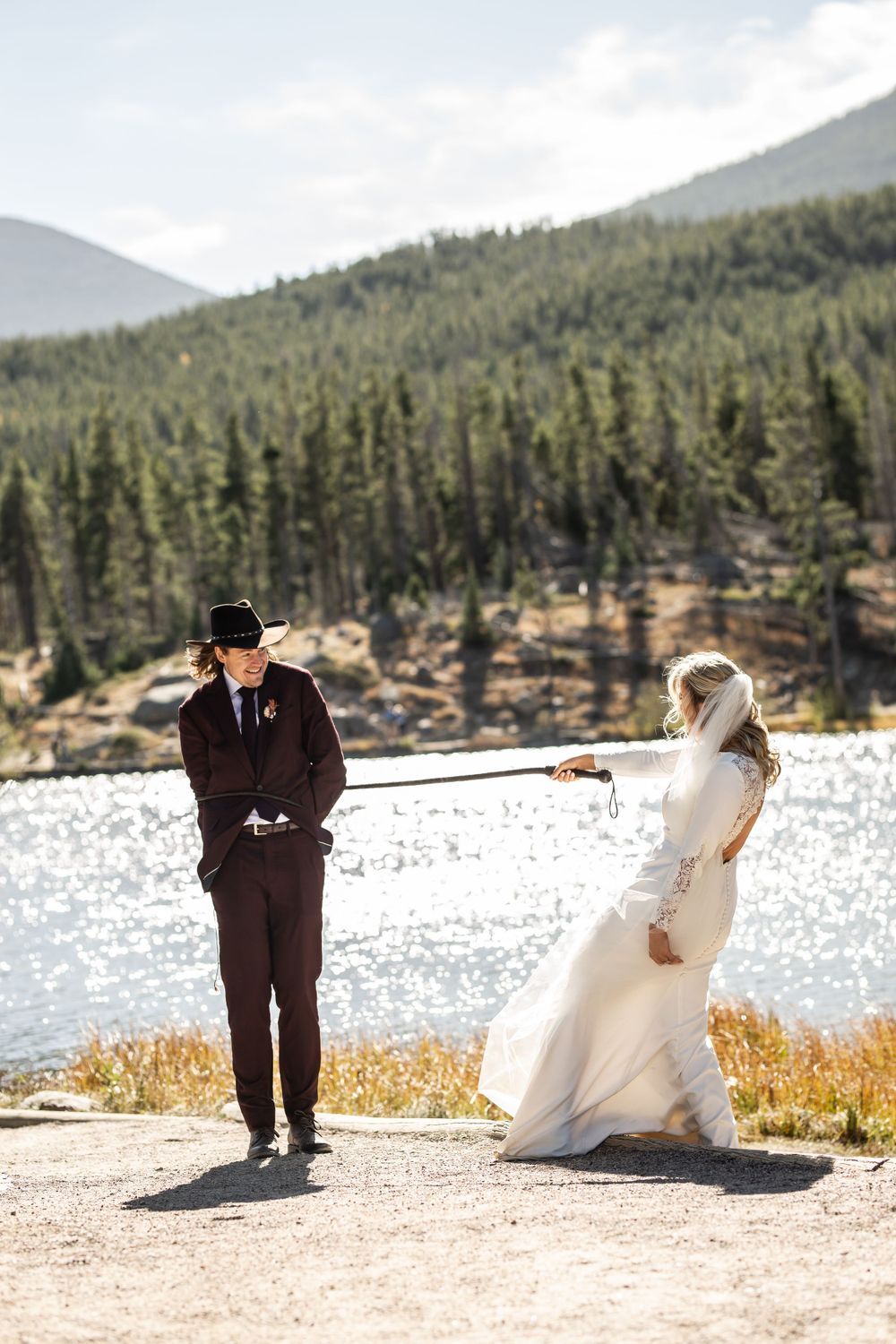 A couple in wedding attire stand by a scenic mountain lake with evergreen trees and sunlit water in the background.