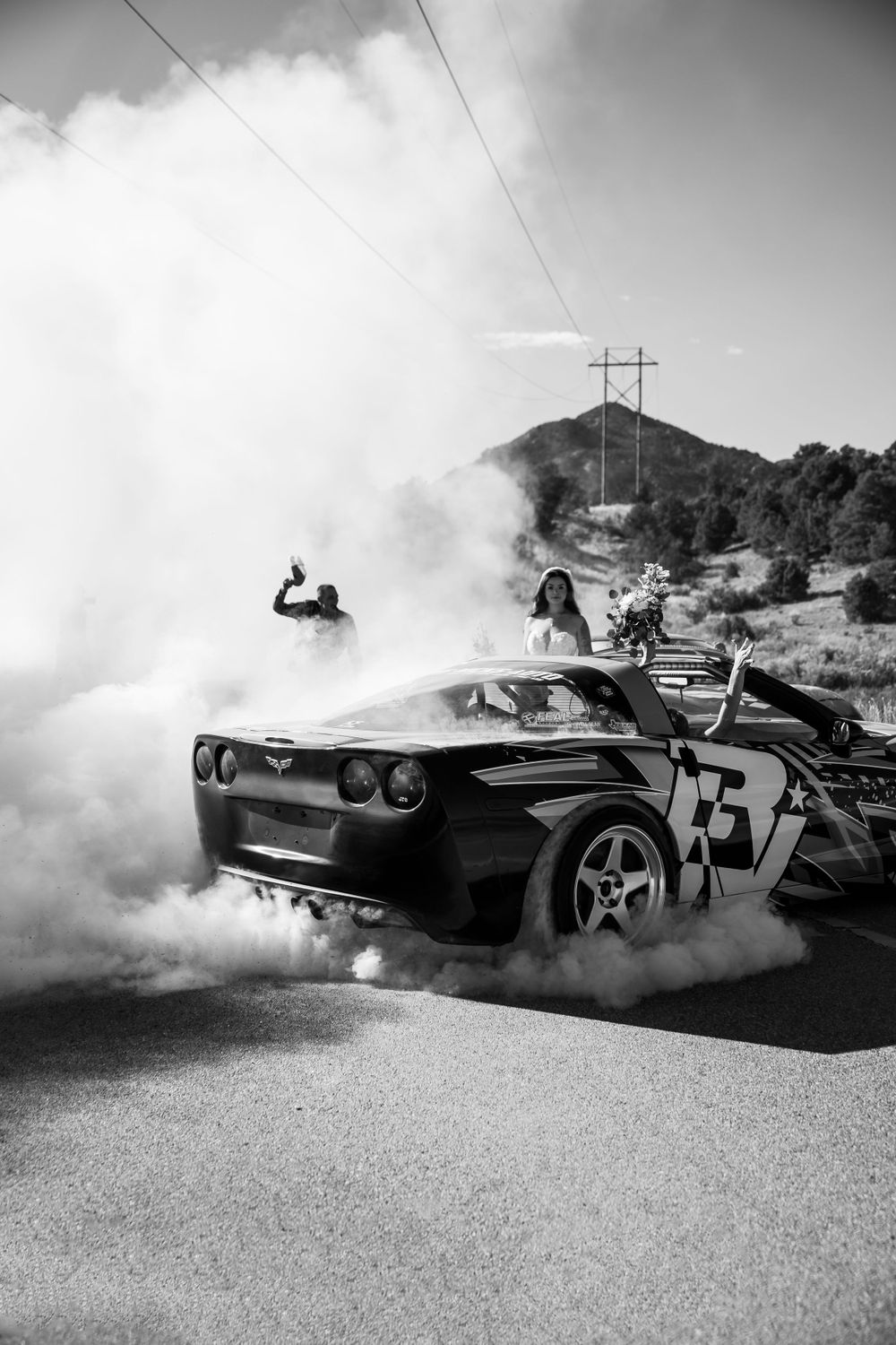 A race car performs a burnout on a mountain road creating dramatic smoke in black and white.