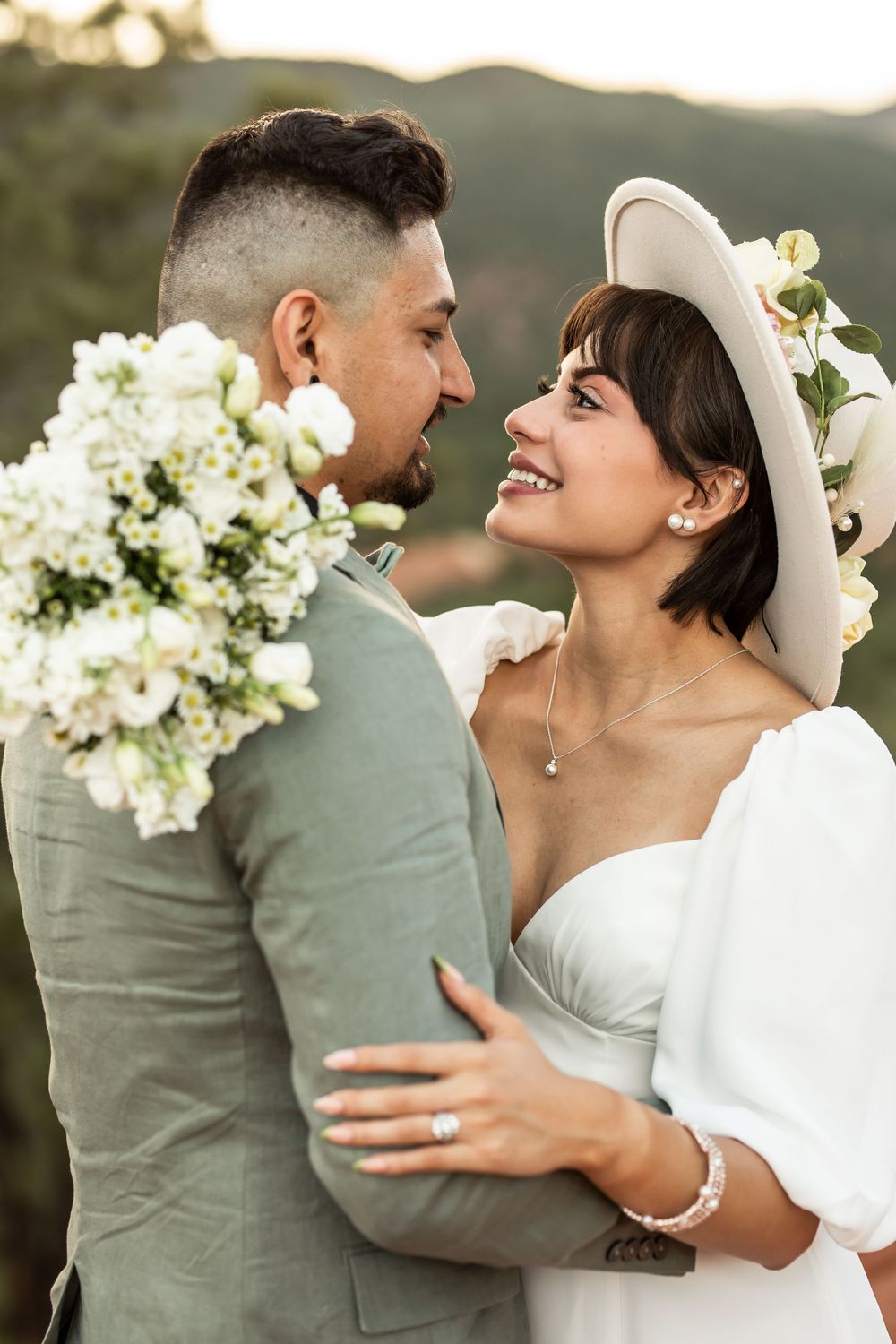 A couple shares an intimate moment outdoors with white floral bouquet and bride wearing a stylish hat.