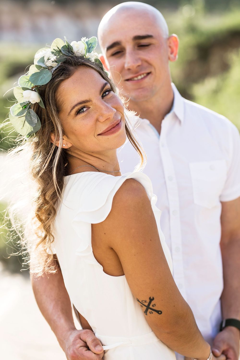 Couple in white clothing poses outdoors, with a floral crown adorning her hair and a small tattoo visible on her arm.