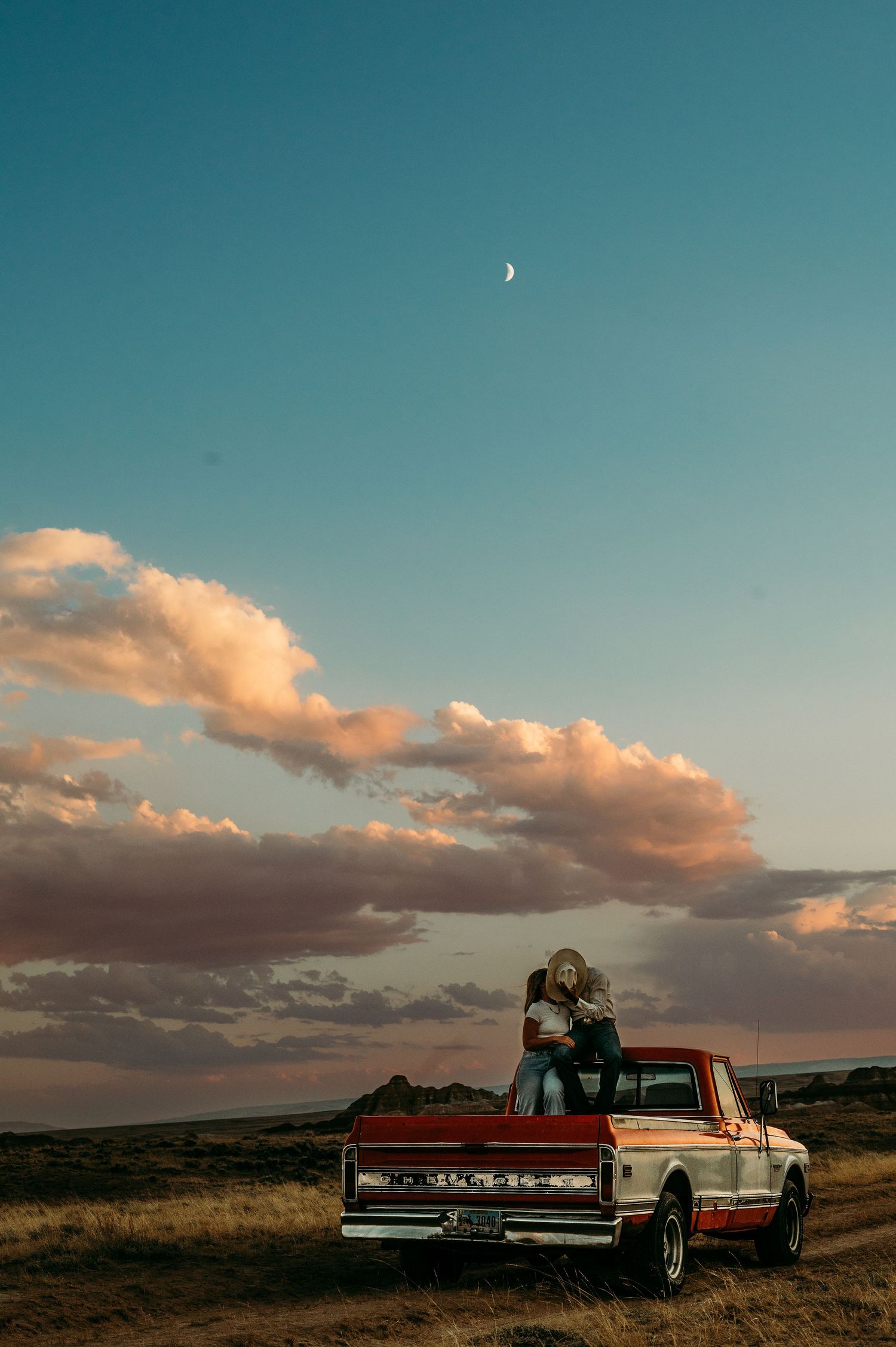Vintage pickup truck under sunset sky with crescent moon and pink clouds.