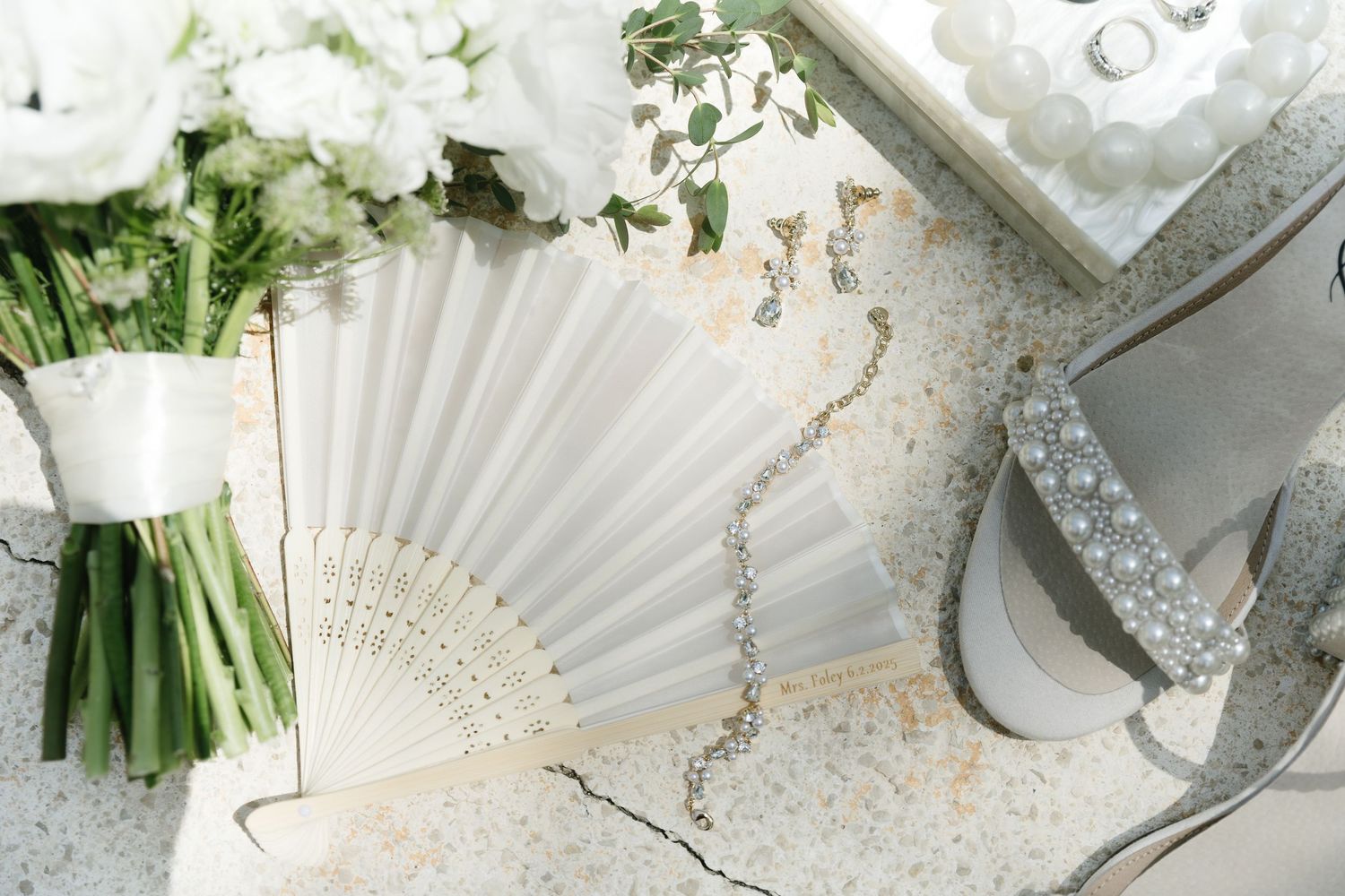 Elegant bridal accessories including white hand fan, sparkly heels, and fresh white floral bouquet arranged on white surface. mexico