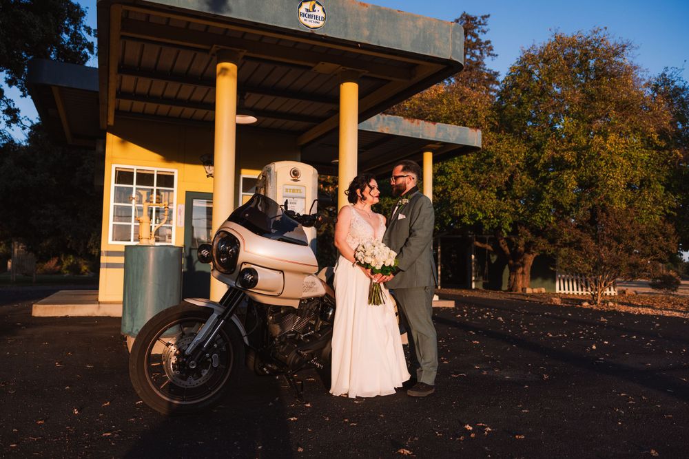 A couple poses with a vintage motorcycle in front of a rustic gazebo during a wedding photoshoot at sunset.