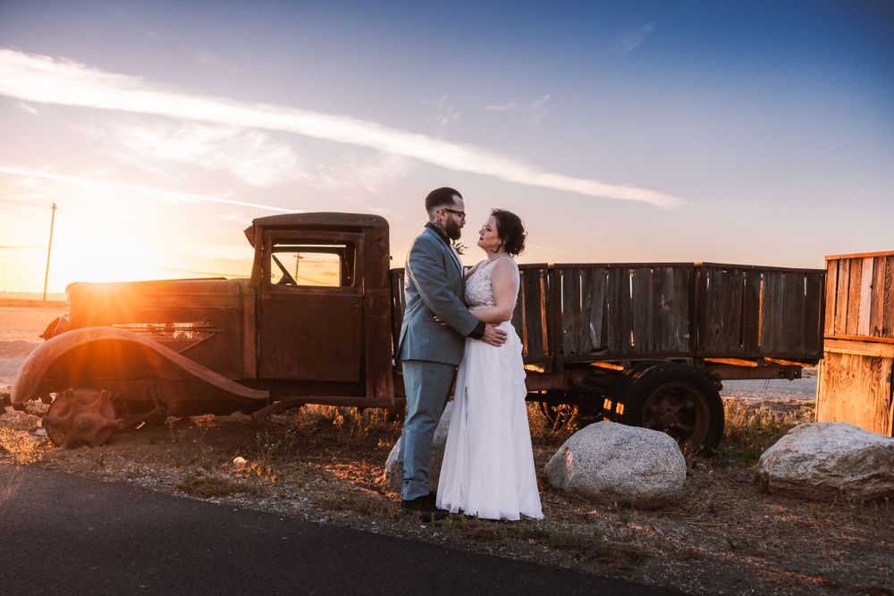 Newlyweds embrace in front of an antique truck during golden hour sunset.