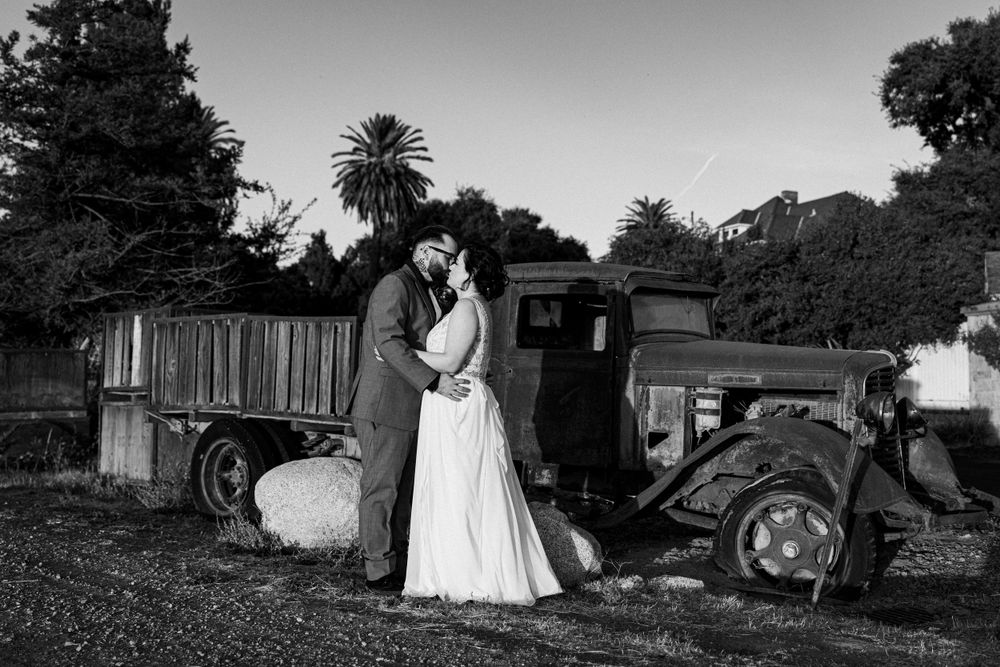 A black and white wedding photo of a couple posing with a vintage car.