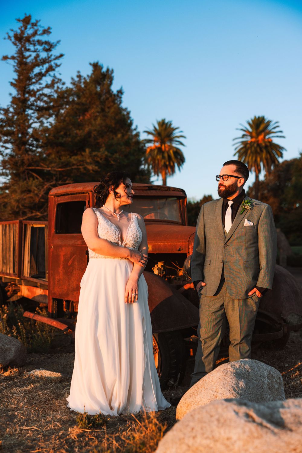 Wedding couple shares romantic moment beside rustic truck with palm trees at sunset.