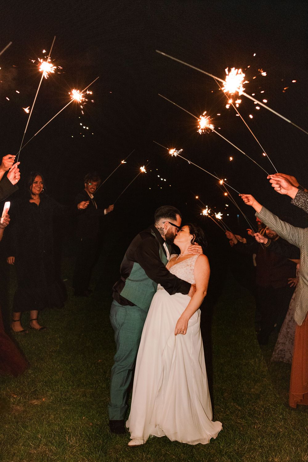 Wedding couple shares intimate kiss surrounded by sparklers held by guests at night.
