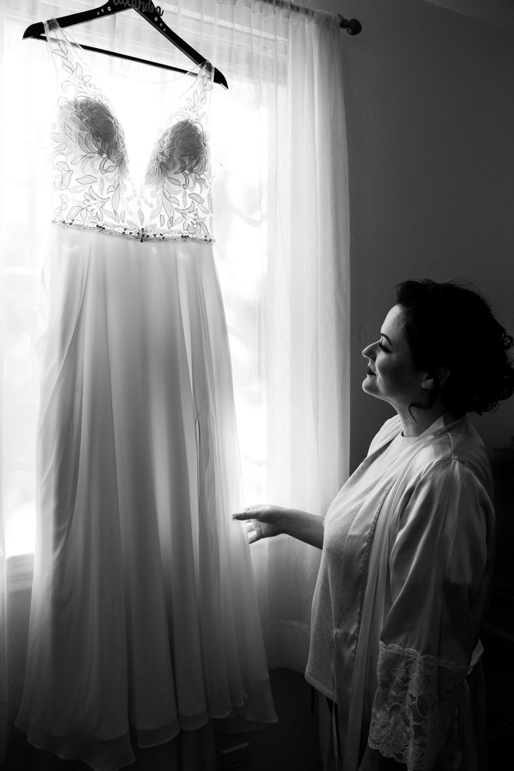 A wedding dress hangs in silhouette against window light as someone gazes up at it.