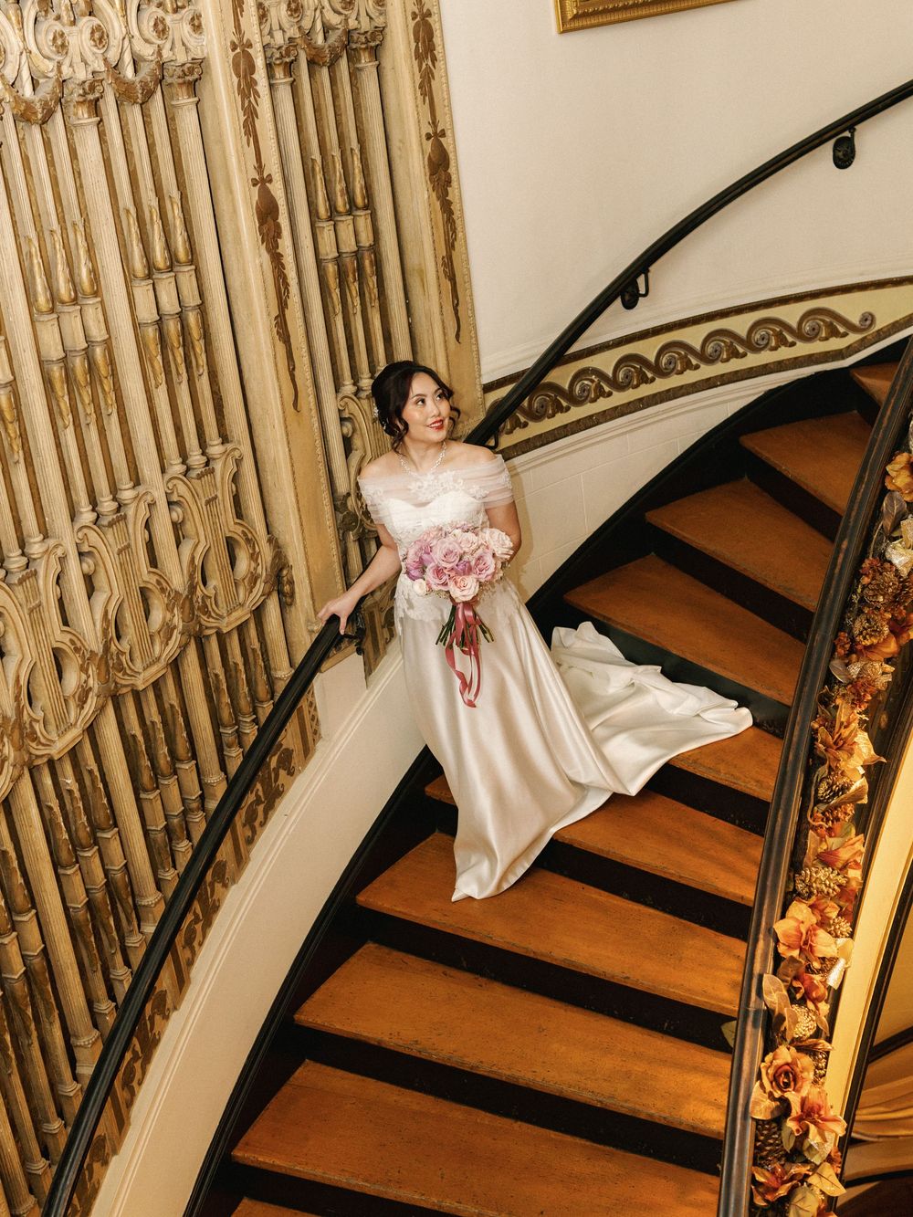 Elegant white wedding dress descending curved wooden staircase with ornate iron railing and floral decor.