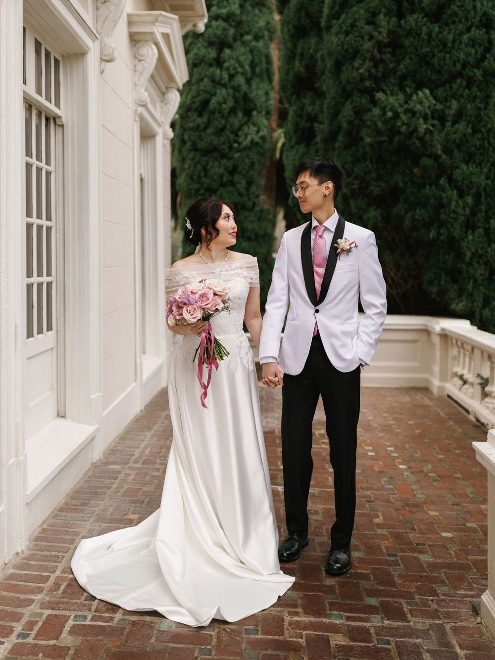 Bride and groom walk together on brick pathway surrounded by white columns and greenery.