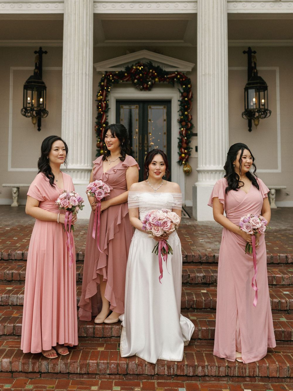 A bridal party in dusty rose and white dresses poses together on brick steps of a classic colonial building.