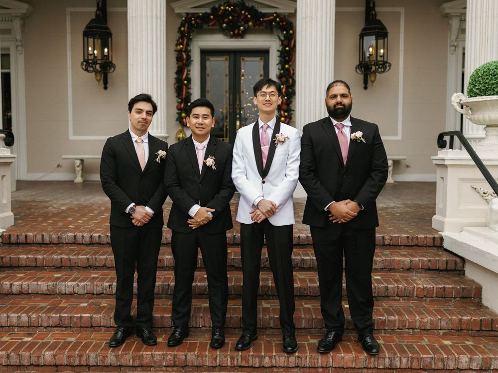 Groomsmen in black suits sitting on brick steps of a white columned building for formal wedding photos.